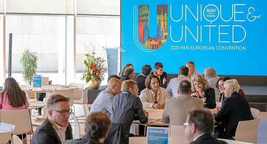 Teilnehmer einer internationalen Tagung sitzen in Gruppen an Tischen und diskutieren, im Hintergrund große blaue Wand mit der Aufschrift Unique & United 2025 Pan European Convention