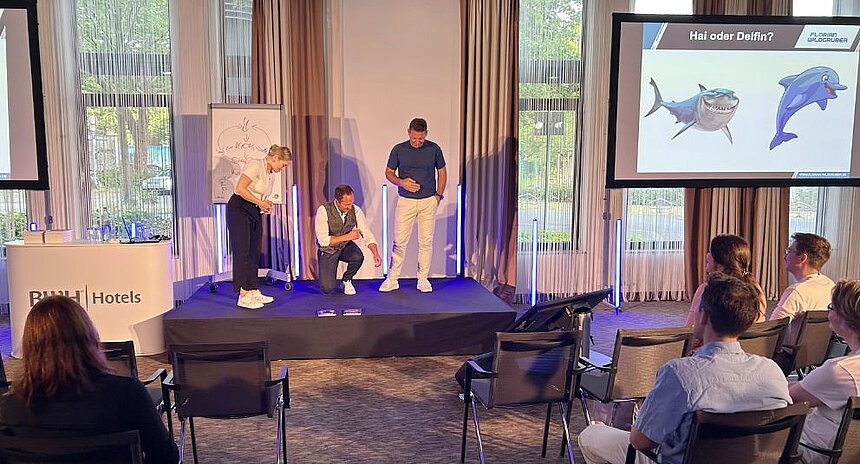 Drei Personen stehen und sitzen auf einer Bühne während einer interaktiven Präsentation, im Hintergrund ist eine Folie mit den Bildern eines Hais und eines Delfins zu sehen, Publikum schaut zu