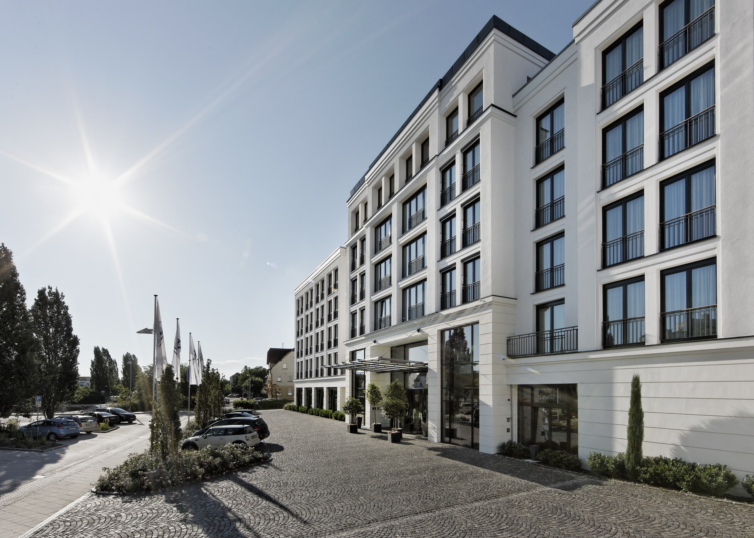 Modernes Stadthotel mit stilvoller Architektur Elegante Hotel-Fassade in zeitloser Architektur mit großen Fenstern und heller Steinverkleidung. Die weitläufige, gepflasterte Eingangszone mit gepflegten Pflanzen verleiht dem Gebäude ein einladendes Ambiente. Strahlender Sonnenschein unterstreicht die moderne und freundliche Atmosphäre des Hotels